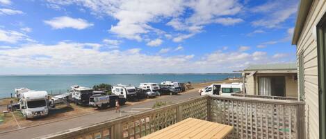 A deck with a wooden table overlooking a parking area with RVs and a view of the ocean.