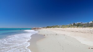 A beach with clear blue water, sandy shore, and buildings in the background.