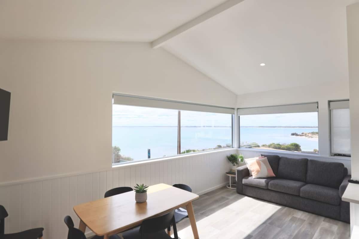 Amplia habitación con un ventanal que ofrece vistas al mar. Hay una mesa de madera con sillas y un sofá gris.