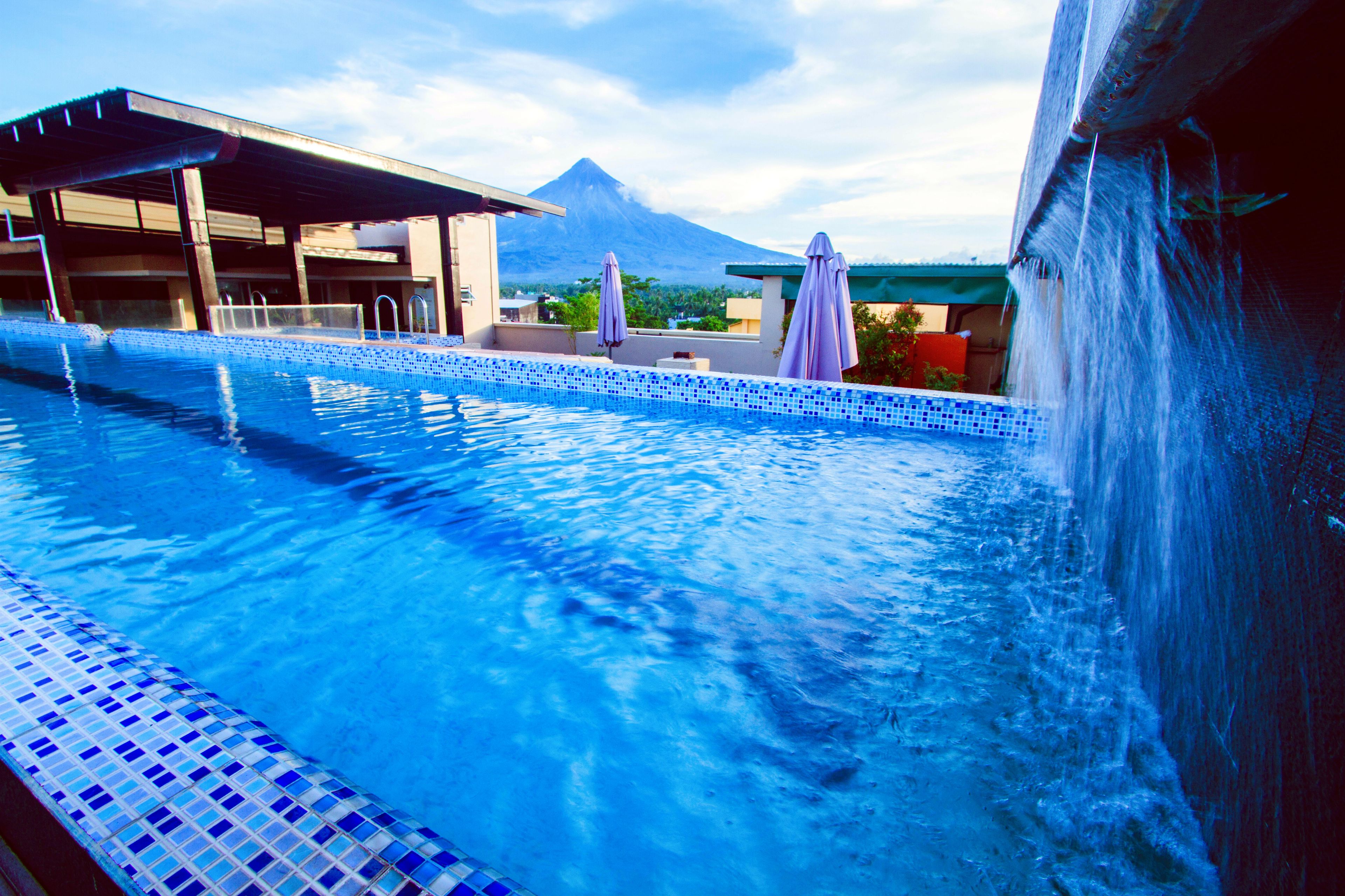 A swimming pool with a tiled edge, a covered area with umbrellas, and a mountain in the background.