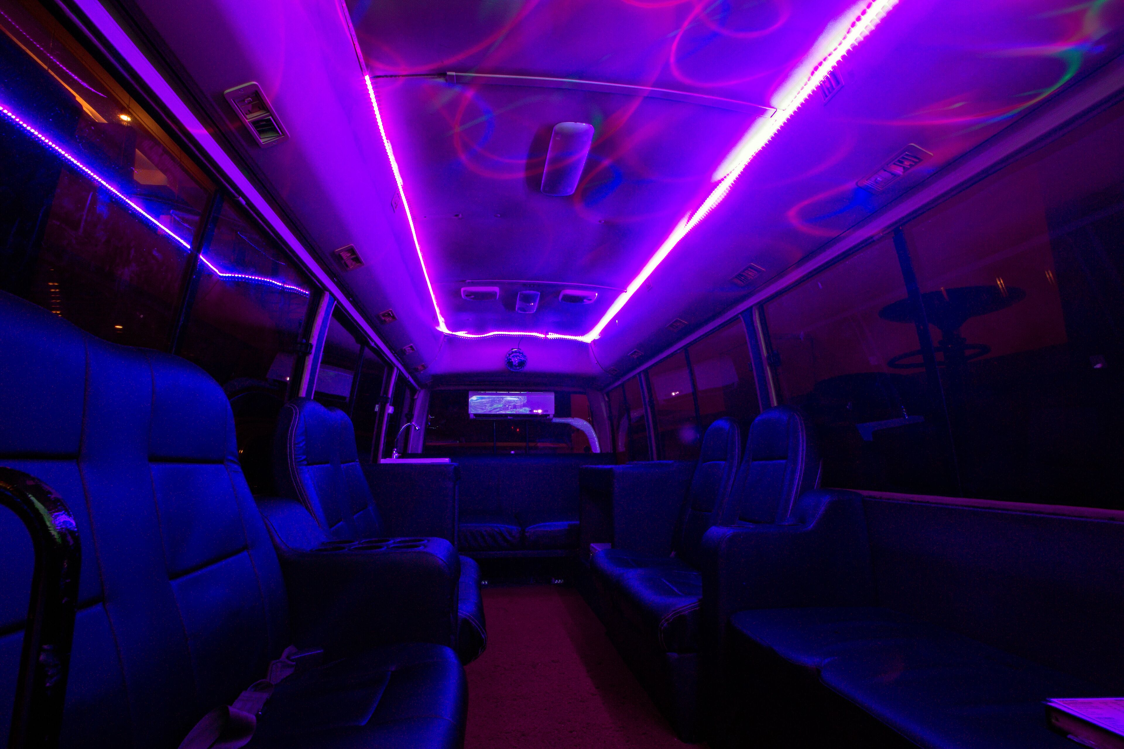 A bus interior with blue seating, a central aisle, overhead lighting, and a television screen.