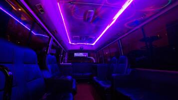 A bus interior with blue seating, a central aisle, overhead lighting, and a television screen.