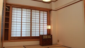 Una habitación japonesa tradicional con suelo de tatami, ventana con celosía de shoji y un pequeño armario de madera con una lámpara.