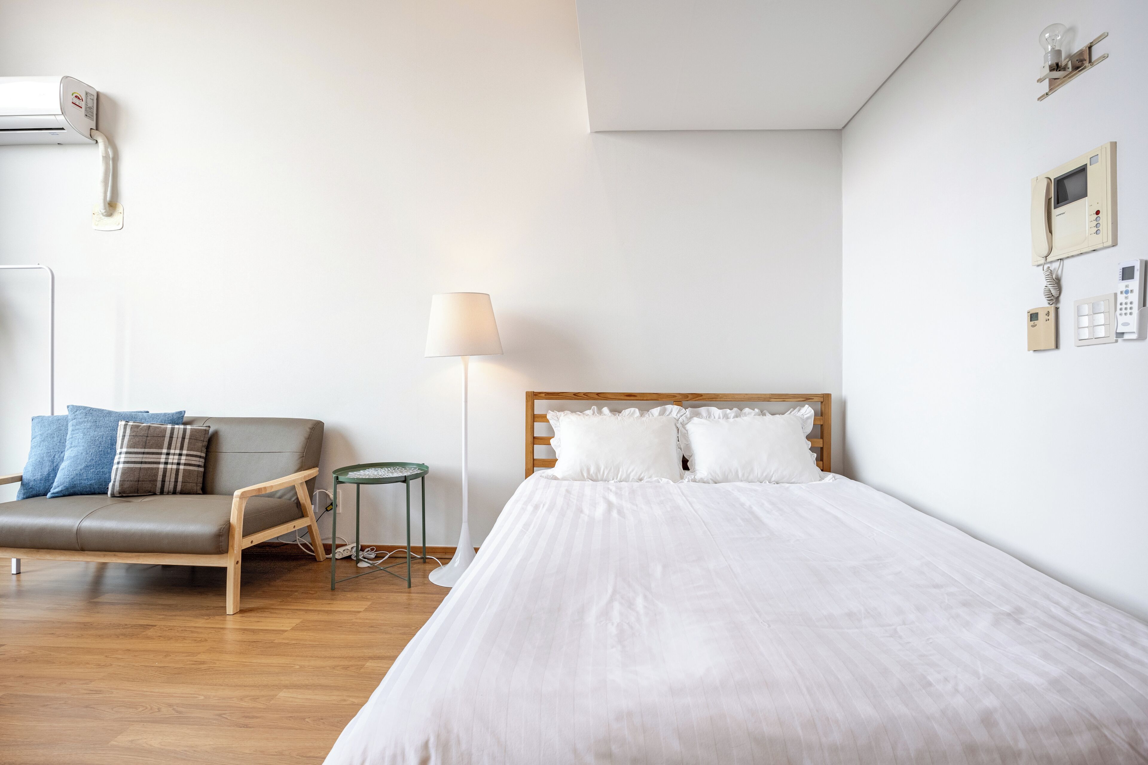 A bedroom with a bed, a wooden headboard, a bedside table, a lamp, a sofa, and a wall-mounted phone.