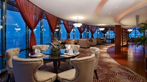 A spacious hotel lobby with a round dining table set for four, large windows with red curtains, and a modern chandelier.