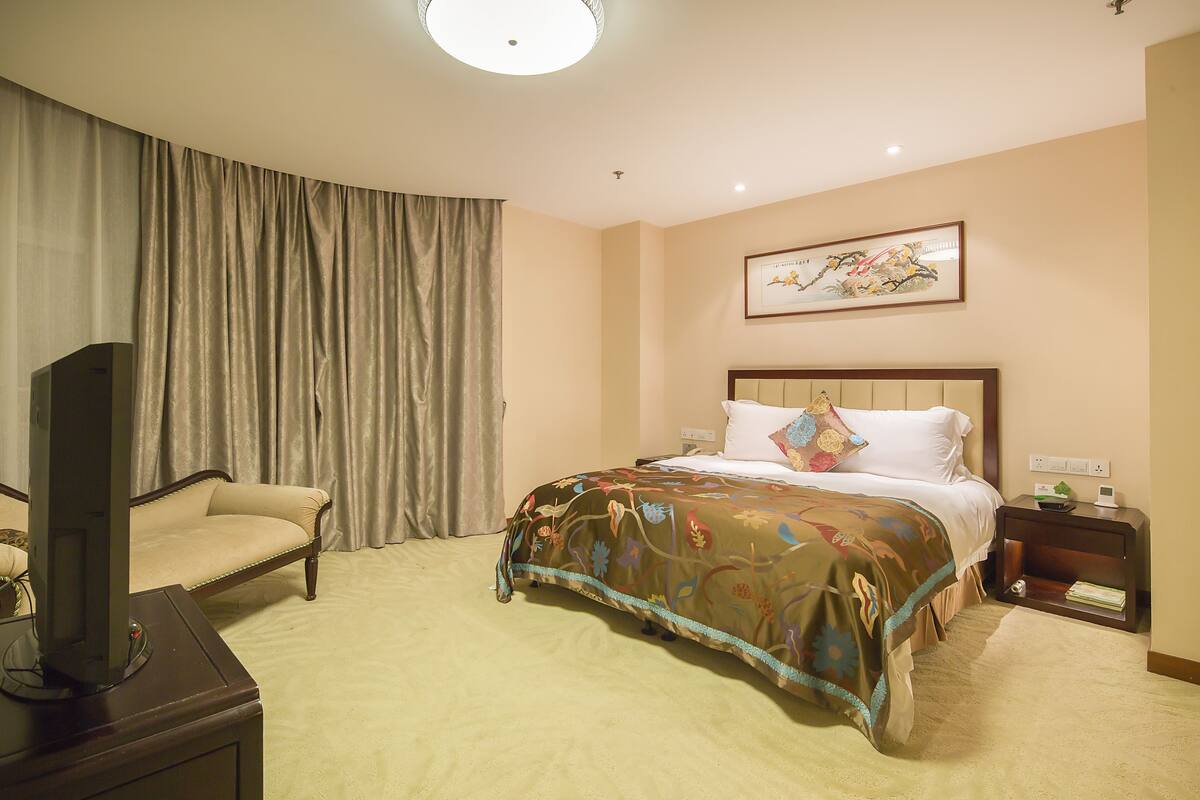 Une chambre d’hôtel avec un lit, un téléviseur posé sur un meuble, une table de chevet avec une lampe et une grande fenêtre avec des rideaux.