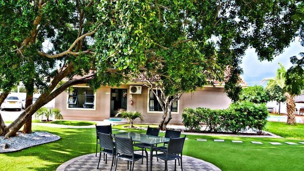 An outdoor patio with a glass table and chairs, surrounded by a well-maintained lawn and a building with a tiled roof. - Cosy Palms Guest House (Gaborone)