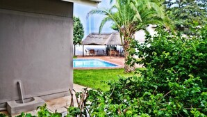 A pool area with a thatched-roof structure, surrounded by greenery and a concrete building. - Cosy Palms Guest House (Gaborone)
