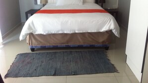 A neatly made bed with a white and red bedspread, a blue and brown skirt, and a grey rug on a tiled floor. - Cosy Palms Guest House (Gaborone)
