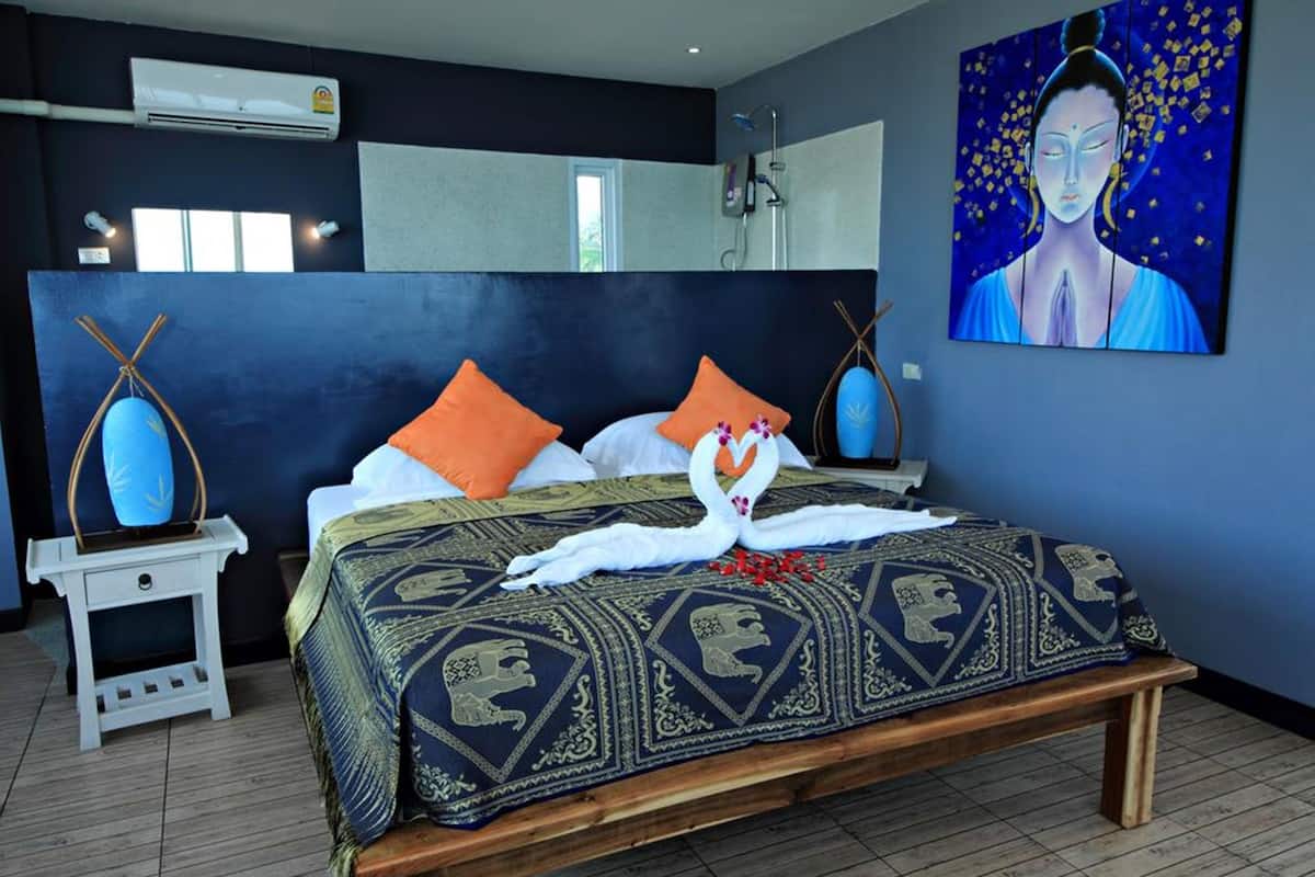 A bedroom with a bed, pillows, a bedside table, a wall-mounted air conditioner, a window, and a painting.
