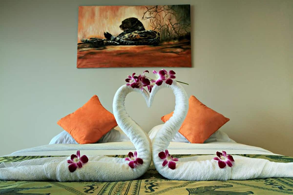 A bed with two towels shaped like swans, orange pillows, and a painting of a dog on the wall.