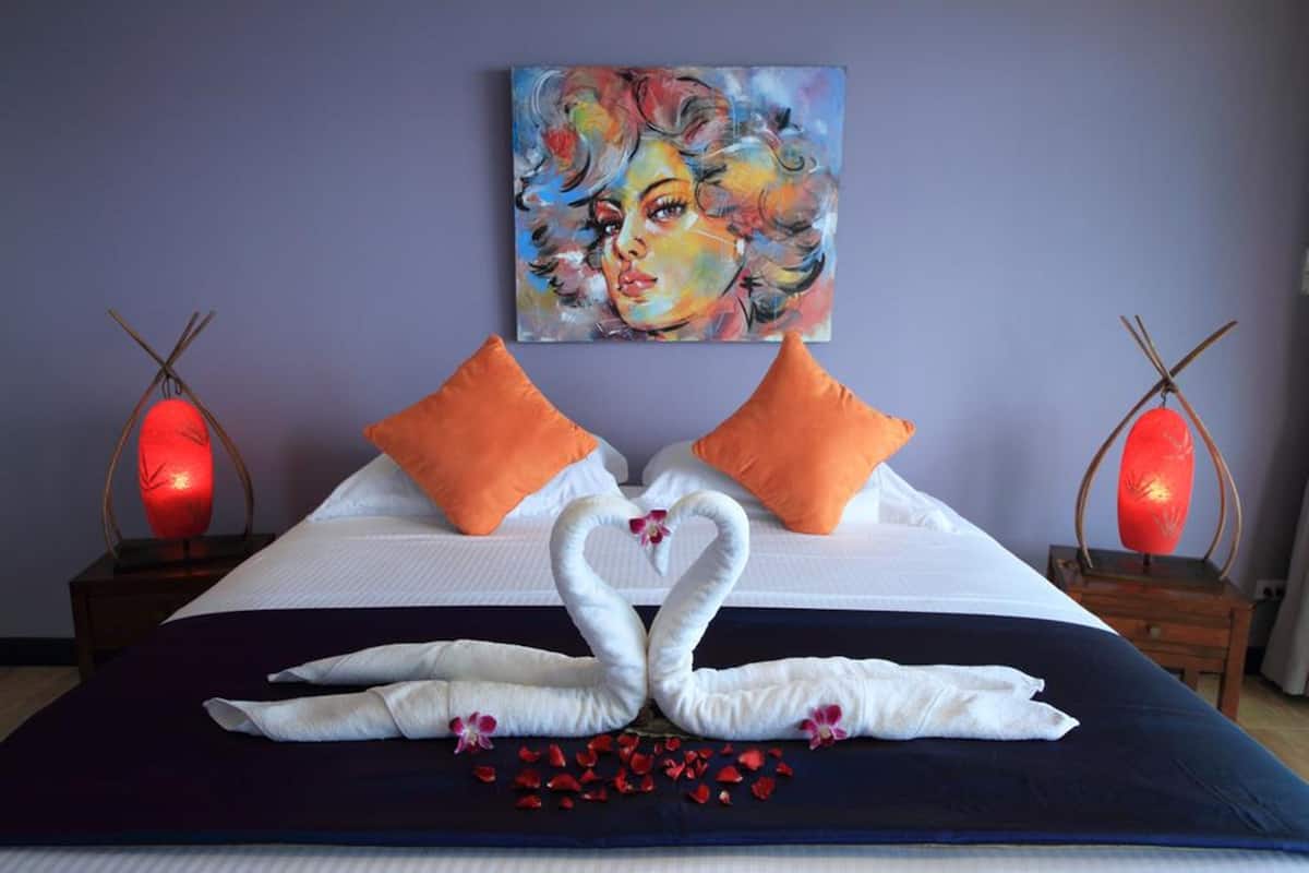 A hotel room with a bed, two orange pillows, a painting on the wall, and two red lamps on nightstands.