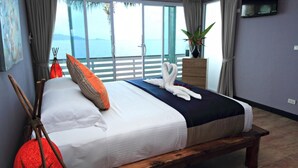 A bedroom with a bed, pillows, a bench, a dresser, a plant, and a TV on the wall. - Anjali (Koh Samui)
