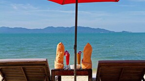 A beach view with two wooden lounge chairs under a red umbrella, a towel, and a drink on a table. - Anjali (Koh Samui)