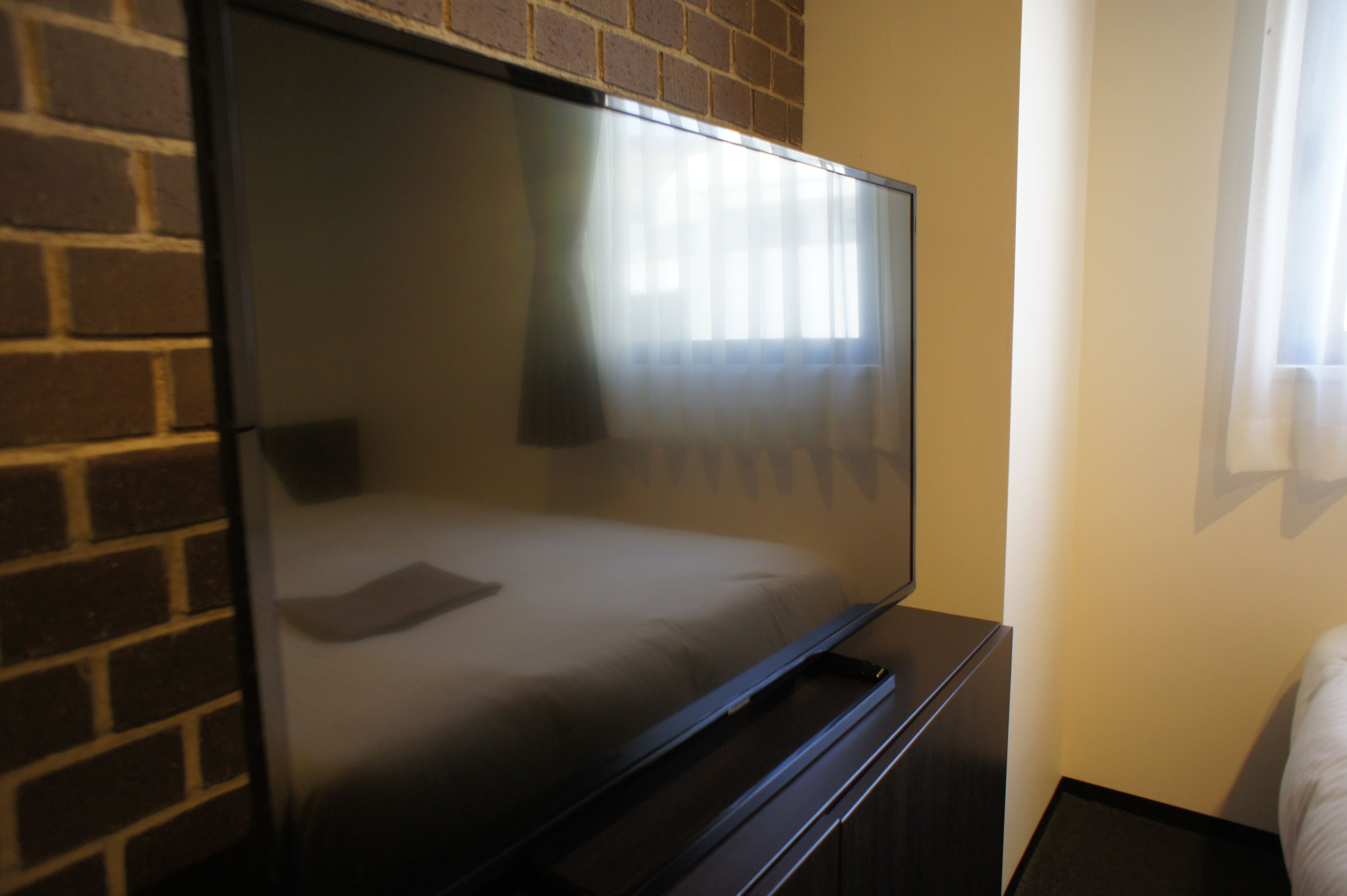 Una habitación de hotel moderna con un televisor de pantalla plana grande montado en una pared de ladrillo, una cama con cabecera oscura y una ventana con cortinas translúcidas.