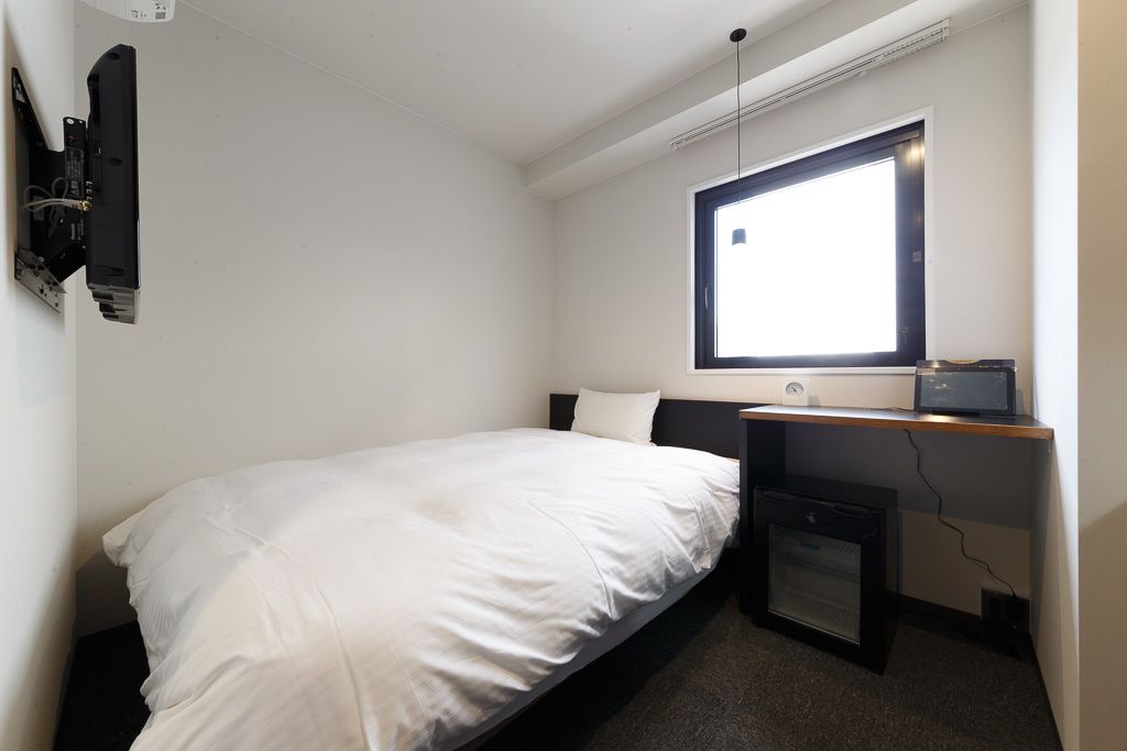 Un dormitorio pequeño y ordenado con una cama individual, un escritorio con computadora y una ventana que proporciona luz natural.