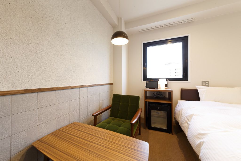 A small hotel room with a single bed, a desk, a chair, a small table, a lamp, and a window.