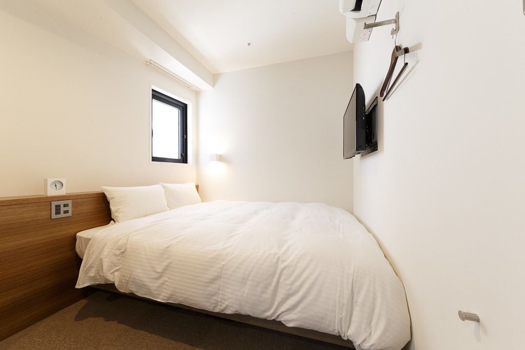 A hotel room with a bed, a window, a wall-mounted TV, and a power outlet.