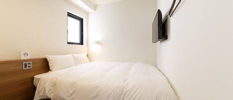 A hotel room with a bed, a window, a wall-mounted TV, and a power outlet.