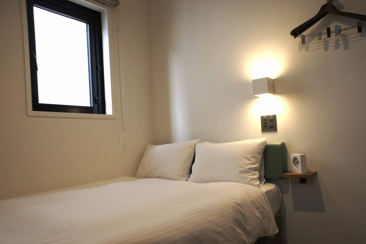 Een net opgemaakt bed in een kamer met een raam, een wandlamp en een kleine speaker op een plank.