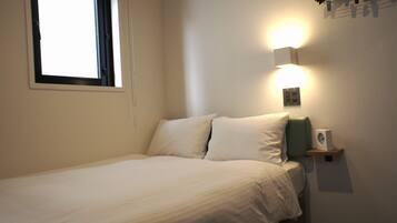 A neatly made bed in a room with a window, a wall-mounted light, and a small speaker on a shelf.