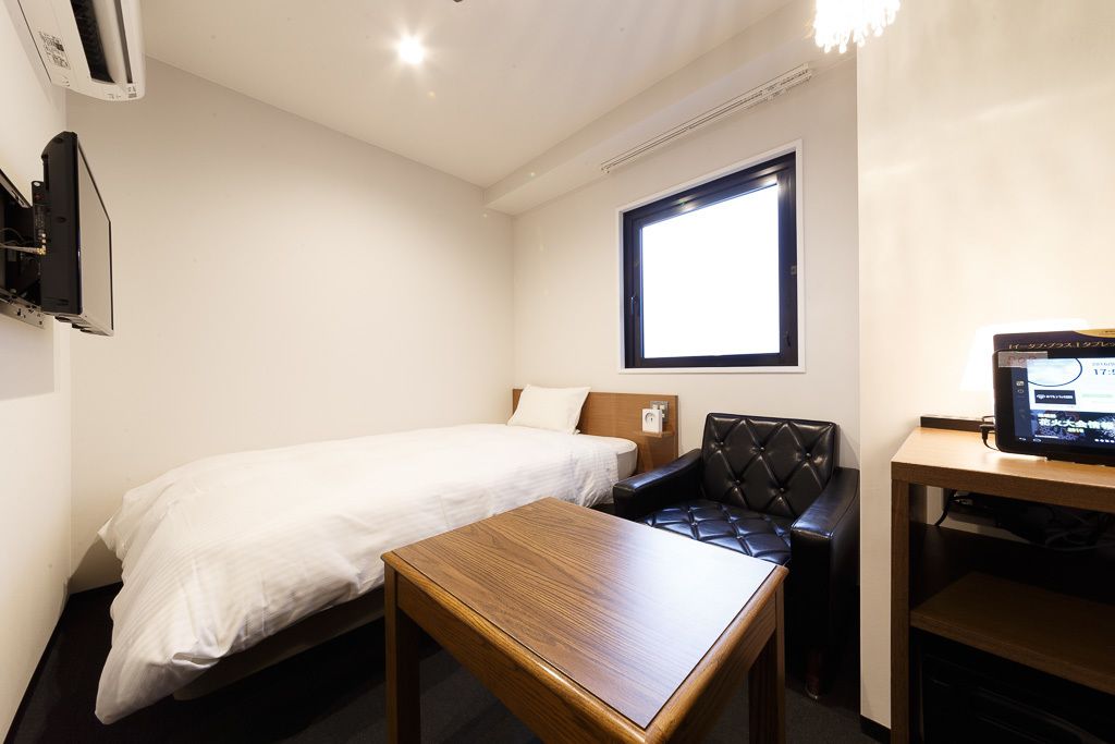 Una habitación de hotel pequeña y ordenada con una cama individual, una mesa de madera, un sillón de cuero, un televisor y una ventana que proporciona luz natural.