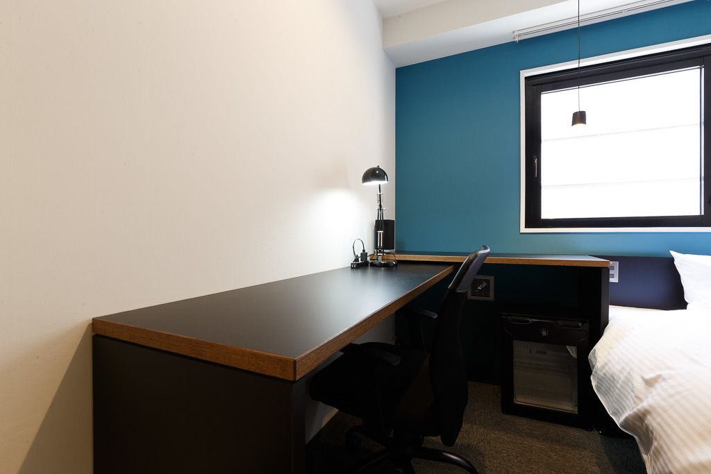 A hotel room with a wooden desk, a black office chair, a small desk lamp, a bed with white bedding, and a window with blinds.