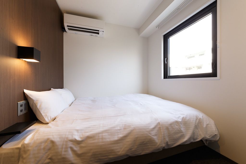 Un lit simple avec un matelas et des oreillers blancs, un climatiseur fixé au mur, une fenêtre donnant sur des immeubles et un luminaire fixé au mur.