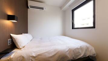Un lit simple avec un matelas et des oreillers blancs, un climatiseur fixé au mur, une fenêtre donnant sur des immeubles et un luminaire fixé au mur.