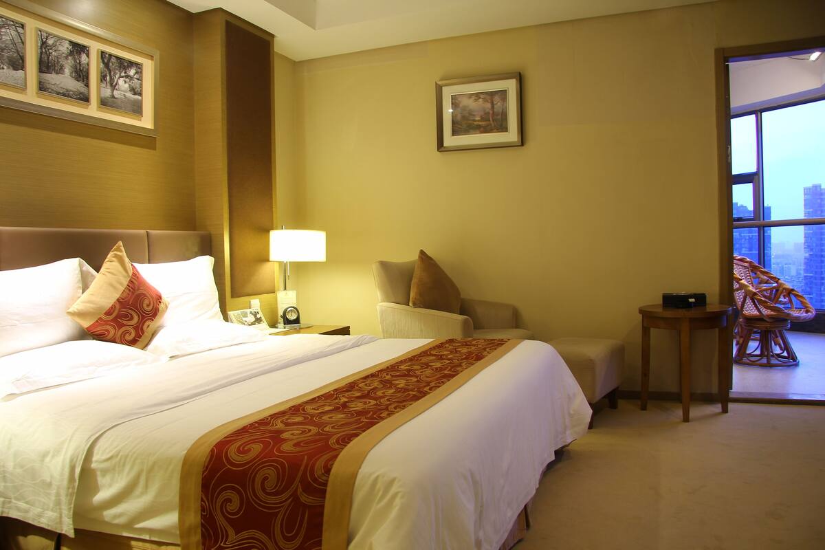A hotel room with a large bed, a nightstand with a lamp, a chair, and a view of the city through a large window.