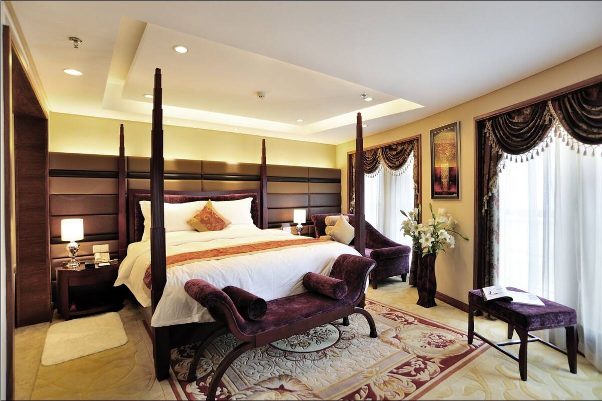 A four-poster bed with a white bedspread and multiple pillows, a nightstand with a lamp, a dark wooden headboard, a large window with curtains, and a decorative rug.