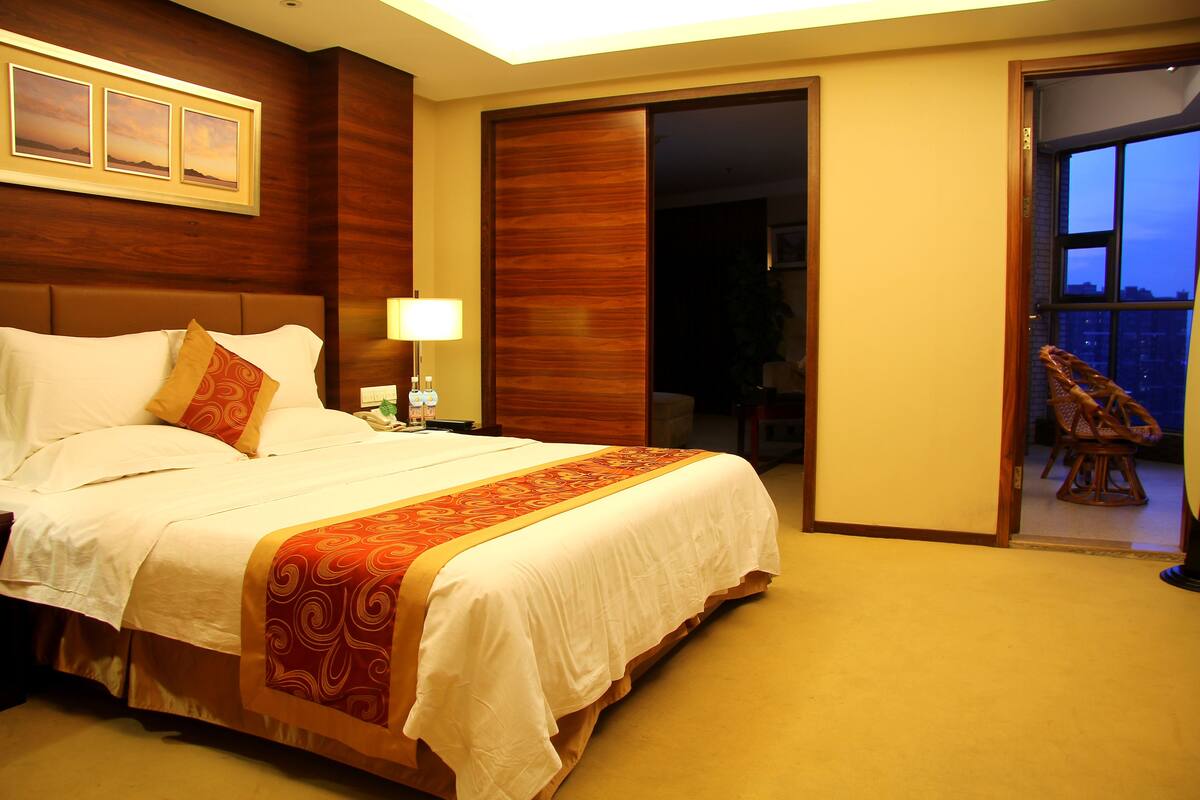A hotel room with a large bed, a nightstand, a lamp, a wooden wardrobe, and a balcony with a chair.