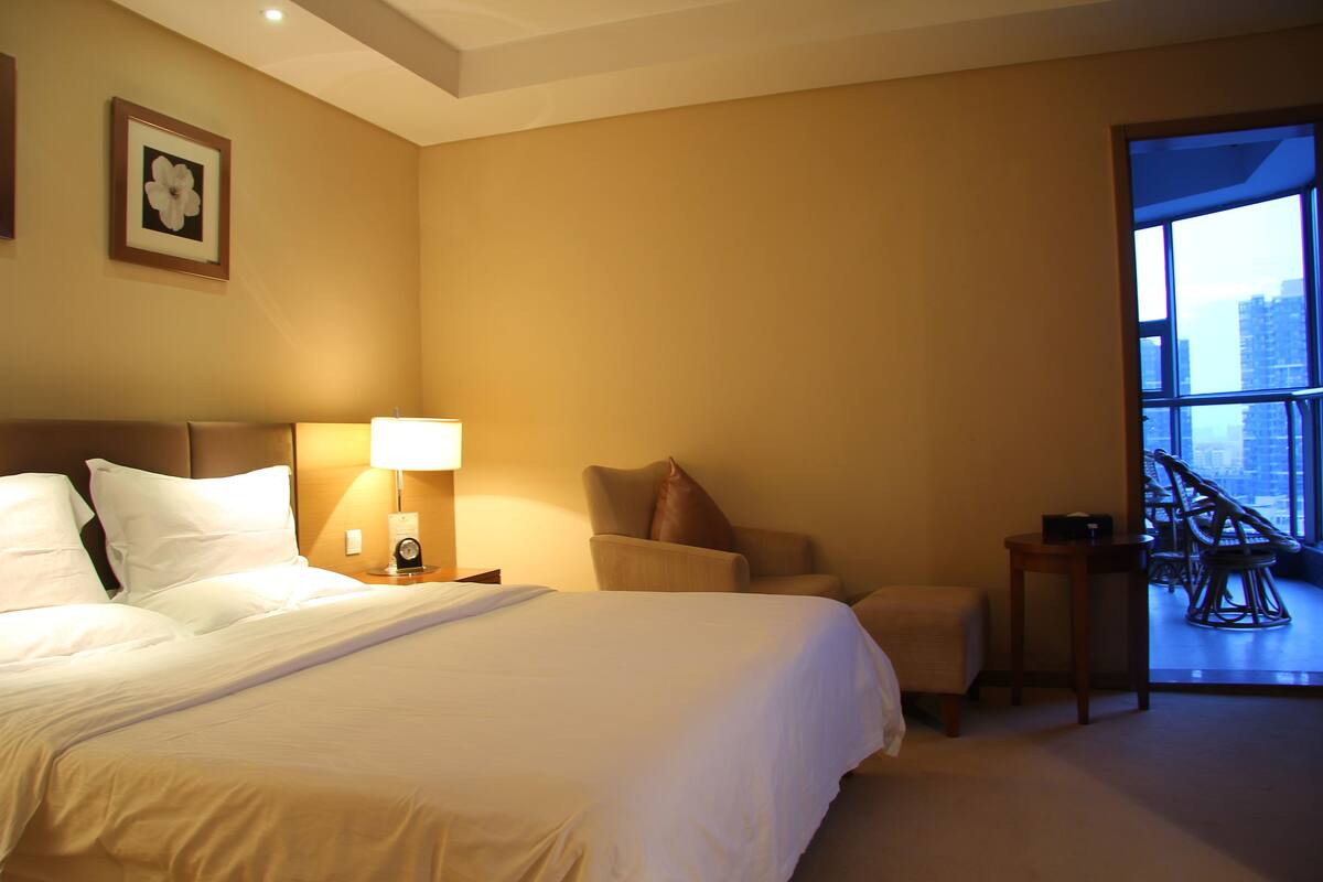 A hotel room with a large bed, a sitting area with a chair and a table, and a balcony with chairs.