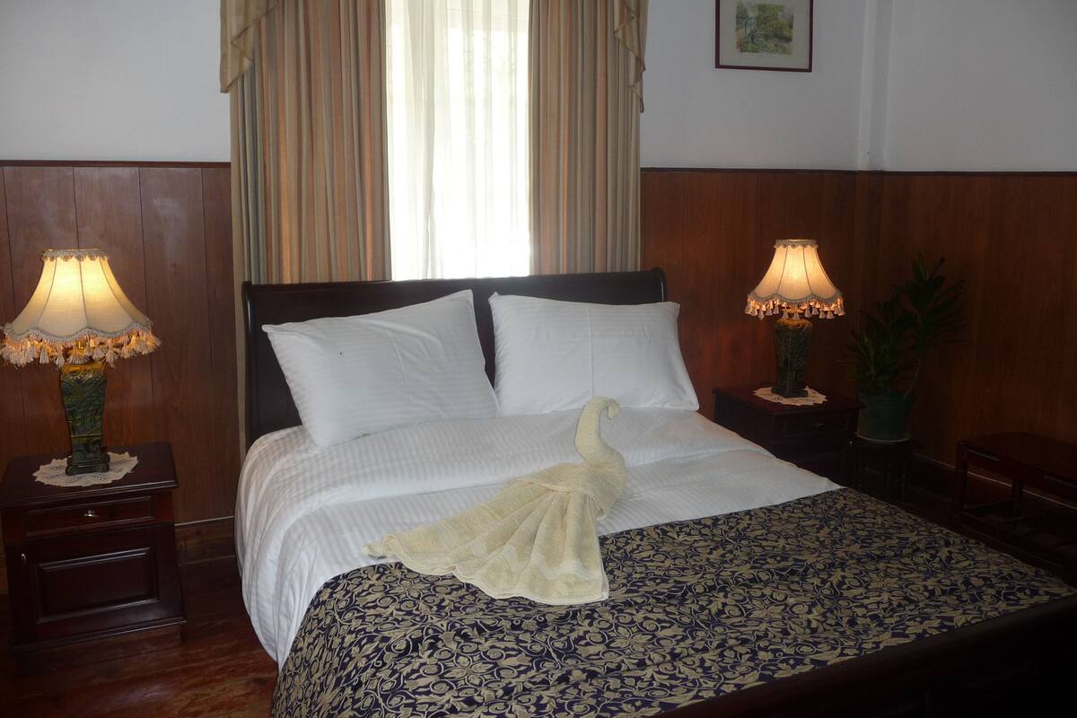 Un lit bien fait, avec du linge de lit blanc et une serviette décorative, encadré par deux tables de chevet avec des lampes, dans une pièce aux murs lambrissés et dotée d’une fenêtre avec des rideaux.