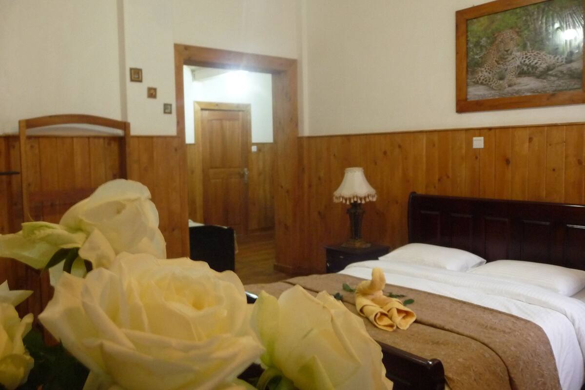 Une chambre d’hôtel avec des boiseries, un lit recouvert de draps blancs, un tableau représentant un léopard et un bouquet de fleurs blanches.
