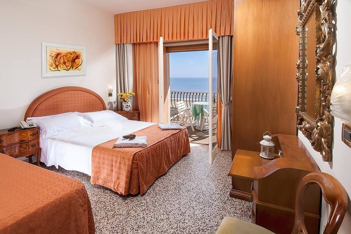 A hotel room with a bed, a desk, a chair, a balcony with a view of the sea, and a painting on the wall.