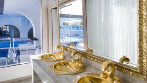 A bathroom with a large mirror, gold fixtures, and a view of a pool and outdoor area. - Dea Hotel (Cervia)