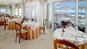 A restaurant with round tables, chairs, and a view of the sea through large windows. - Dea Hotel (Cervia)
