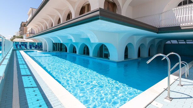 A swimming pool with clear blue water, surrounded by white buildings with arched windows and balconies. - Dea Hotel (Cervia)