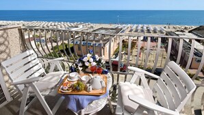 A terrace with white chairs and a table set with a tray of food and drinks, overlooking a beach with umbrellas and people. - Dea Hotel (Cervia)