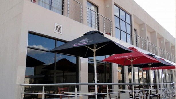 An outdoor seating area with tables and chairs, umbrellas, and a building facade with large windows and a balcony. - Hotel LaBama (Gaborone)