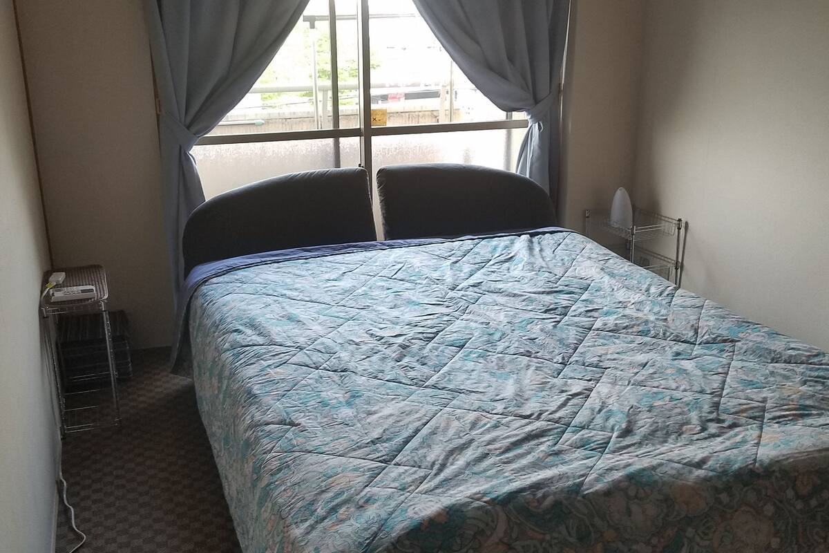 Un dormitorio con cama, ventana con cortinas, unidad de aire acondicionado y una mesita de noche pequeña.