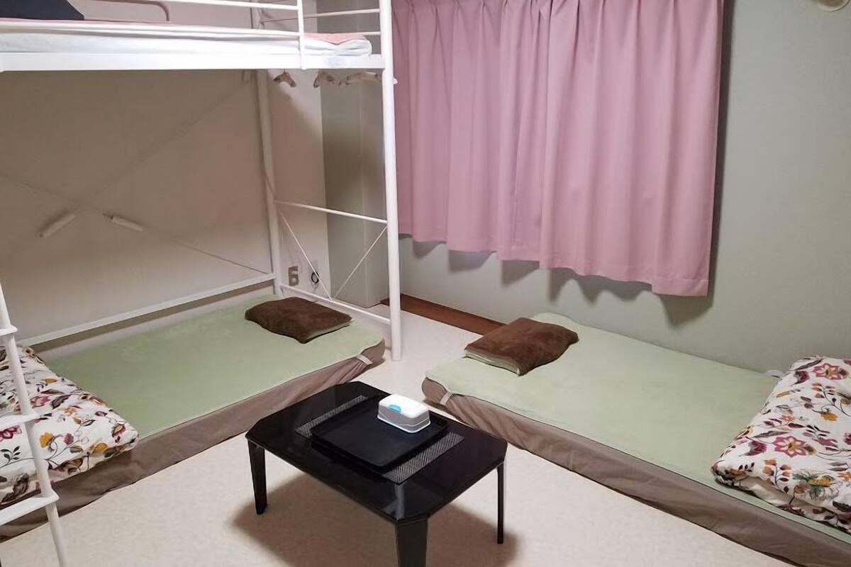 Una habitación pequeña y bien iluminada con una cama nido, una cama individual, una mesita con un dispositivo electrónico y una ventana con cortinas rosadas.
