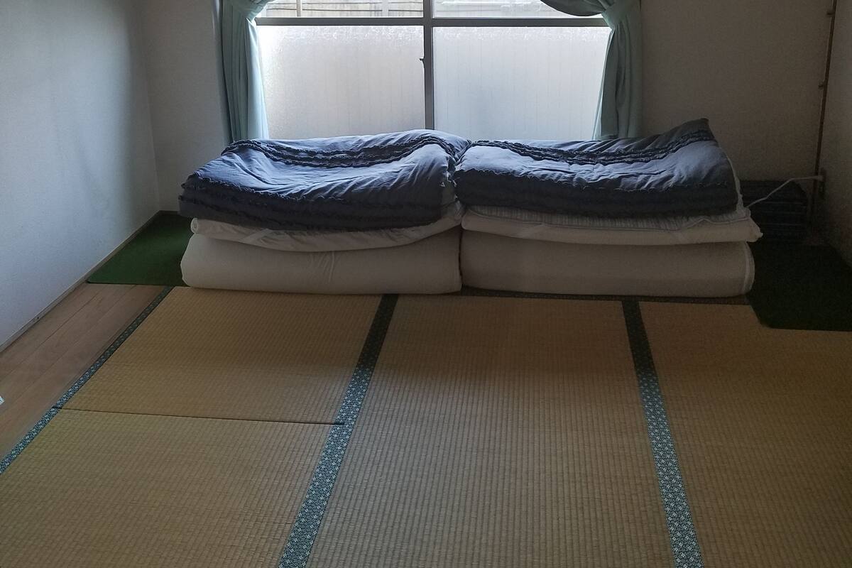 Una habitación con suelo de tatami, una cama con colcha azul y blanca, y una ventana con cortinas verde claro.