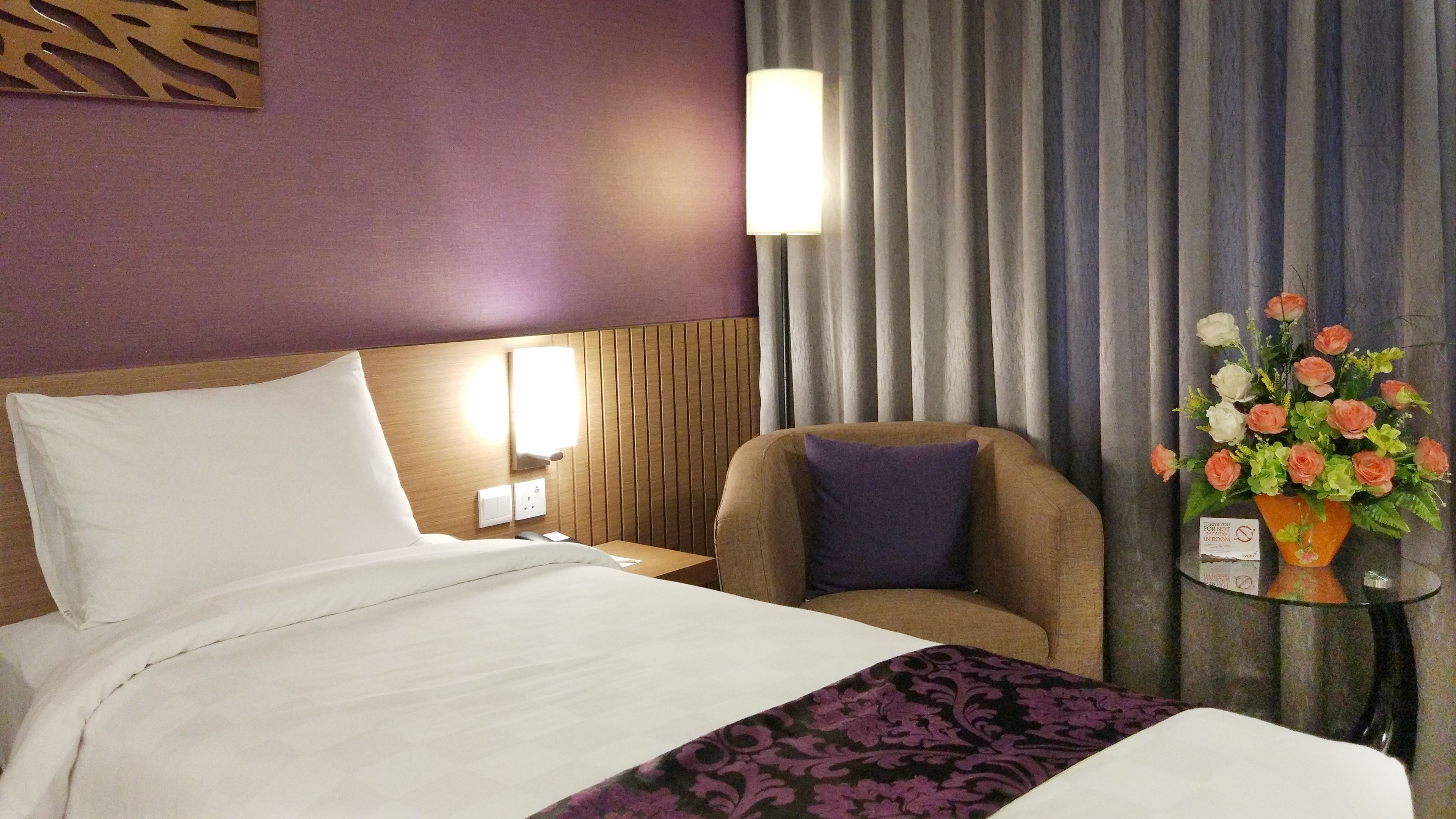Une chambre d’hôtel avec un lit, une chaise, une table de chevet, une lampe et un vase à fleurs.