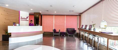 A hotel reception area with a modern design, featuring a reception desk, seating area, and a view of the outdoors through large windows.
