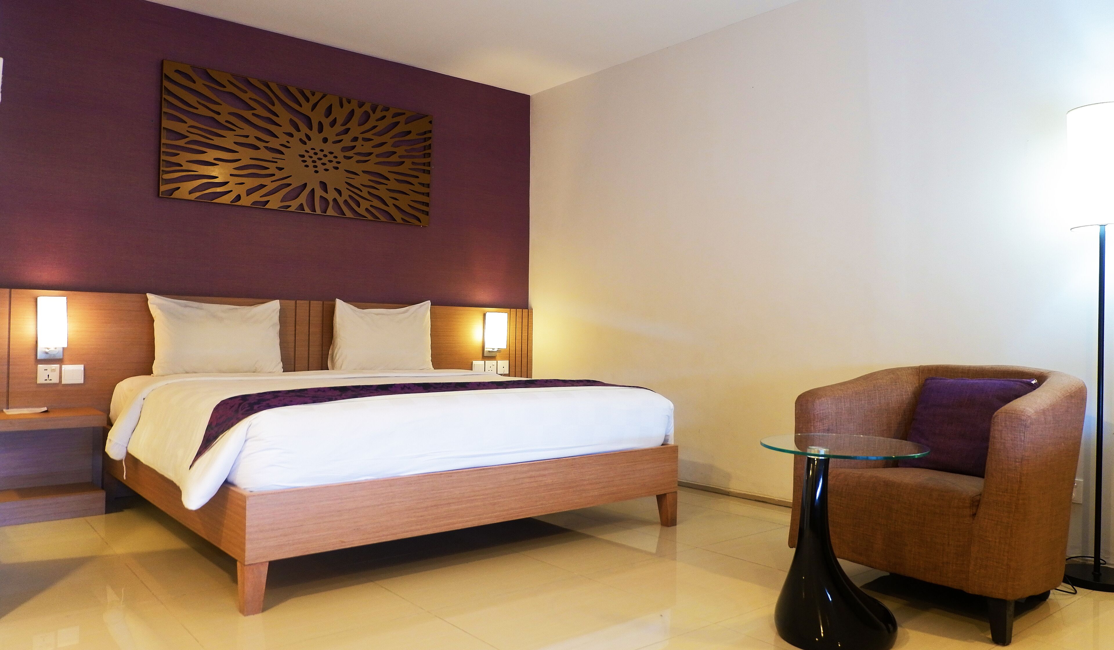A hotel room with a bed, two bedside lamps, a decorative wall piece, a chair, and a floor lamp.