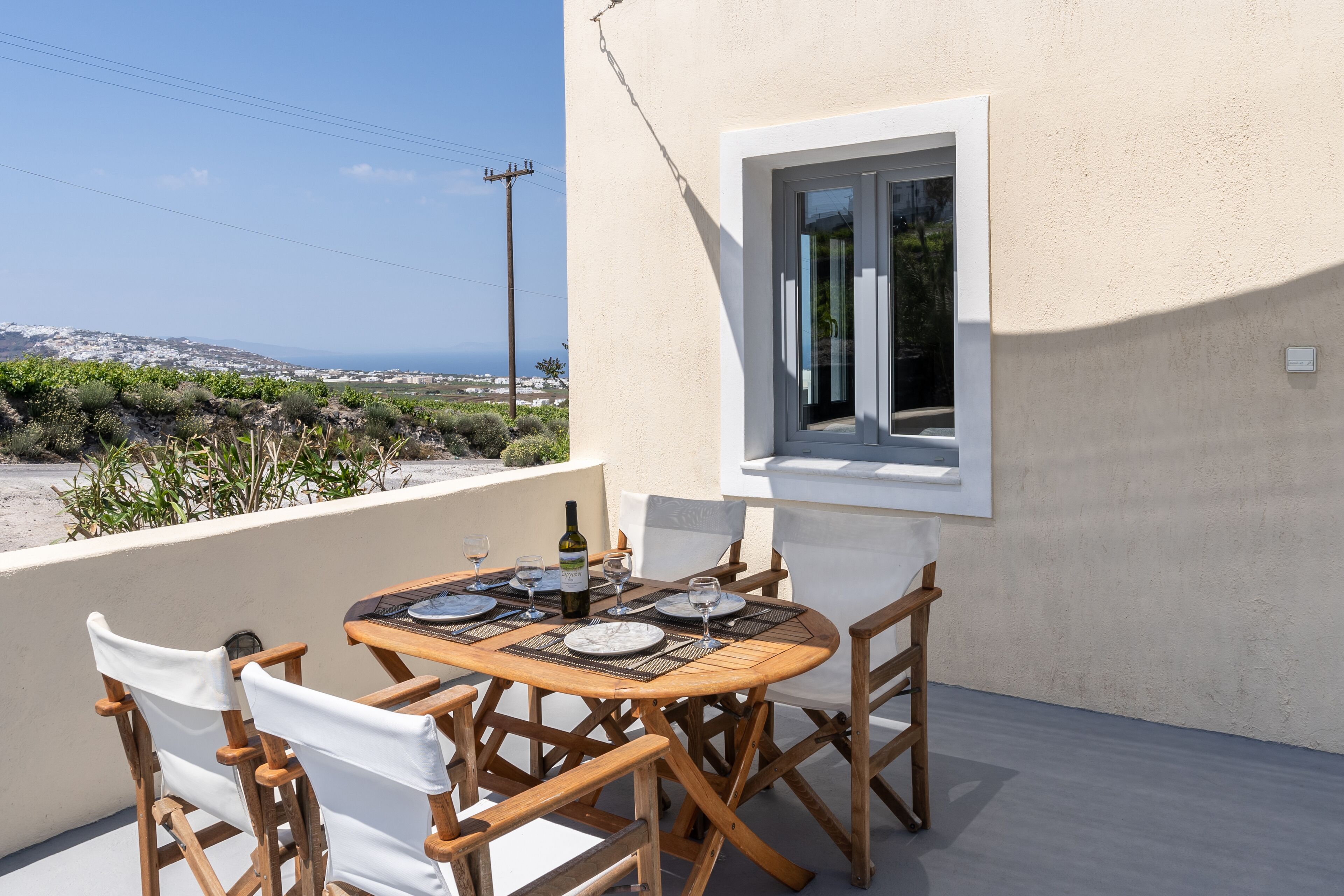 Une table en bois, dressée pour le repas, entourée de chaises blanches, une fenêtre avec des stores et une vue sur la mer et les bâtiments éloignés.