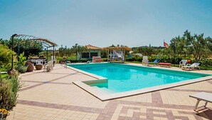 A rectangular swimming pool with a tiled deck, surrounded by lounge chairs and a covered seating area.