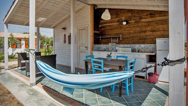 A covered outdoor area with a hammock, blue chairs, a table, and a kitchen area with wooden walls and tiled flooring.
